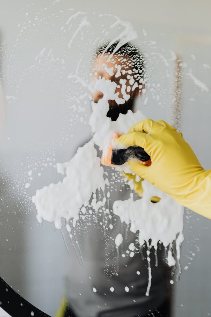 Anonymous male housekeeper in yellow rubber gloves wiping foam from round mirror in bathroom